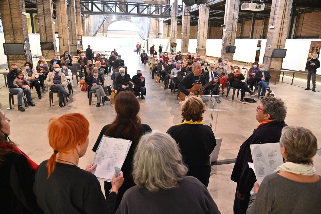 Un coro cantando ante un grupo de personas sentadas en sillas en filas ordenadas en una nave industrial.